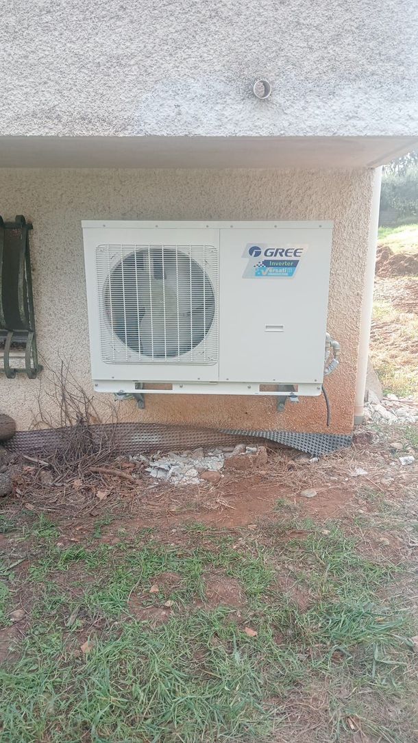 Unità di condizionamento esterna installata sotto un patio coperto, addossata a un muro beige, con erba e pacciame al di sotto.