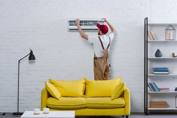 Una persona installa un condizionatore d'aria a parete sopra un divano giallo in un soggiorno moderno.