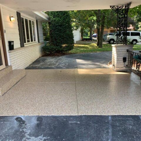 Front porch with textured concrete floor, white brick house, and a shaded driveway with parked cars nearby
