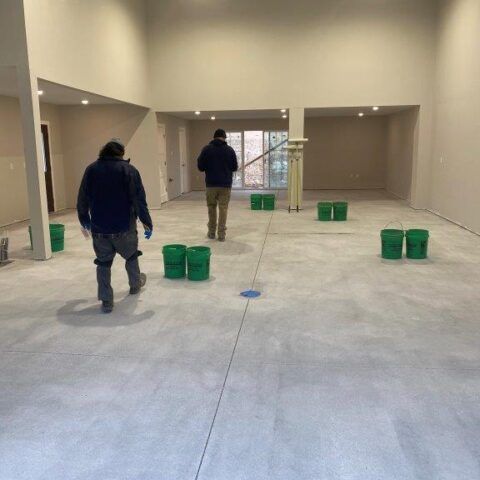 Two workers in a bare room with green buckets on the floor, preparing the space under construction.