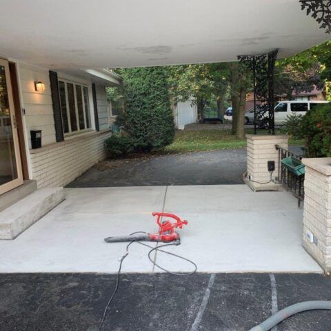 Red power tool on a white driveway under a carport, with a black cord and grassy yard beyond.