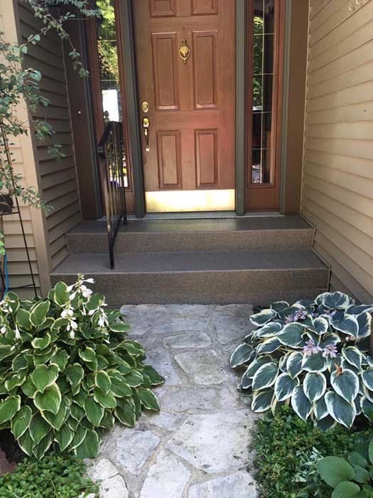 Front porch with a wooden front door, stone walkway, steps, and two leafy potted plants.