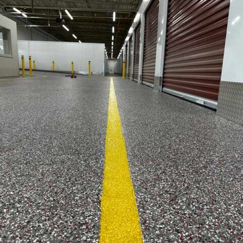 Yellow line on a speckled warehouse floor leading toward storage units and bollards