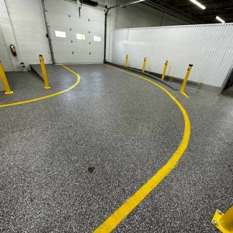 Yellow painted lane curves across a gray speckled warehouse floor with yellow bollards and white walls.