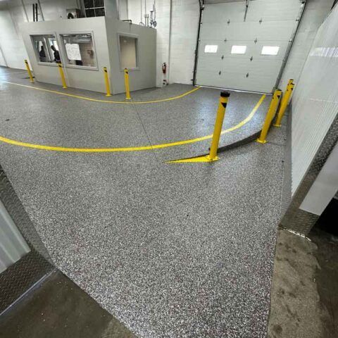 Industrial interior with speckled floor, yellow safety bollards, and large gray garage doors.