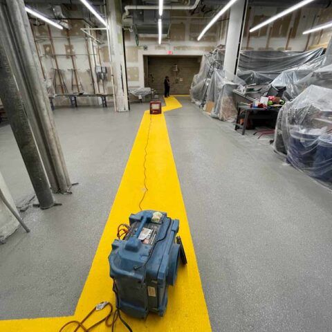 Yellow walkway in an industrial basement with tools and equipment wrapped in plastic on both sides.