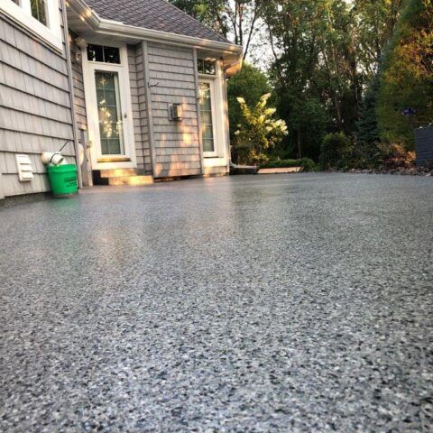 Freshly coated gray driveway beside a house, with sunlight and trees in the background