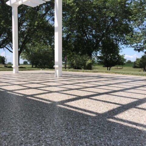 Shaded porch with white columns casting diamond shadows on a gravel patio beside trees and a grassy yard