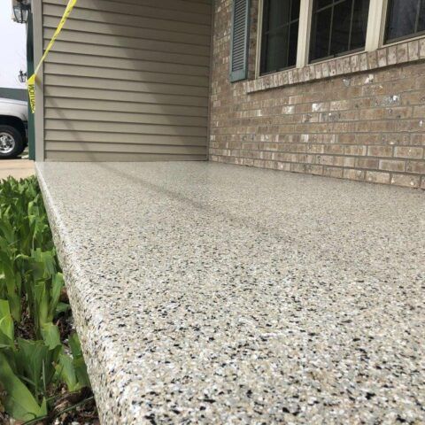 Speckled concrete porch beside tan siding and brick wall with window; green plants at left.