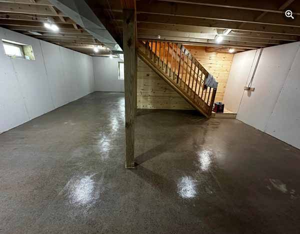 Empty basement with concrete floor, white walls, wooden staircase, and support posts