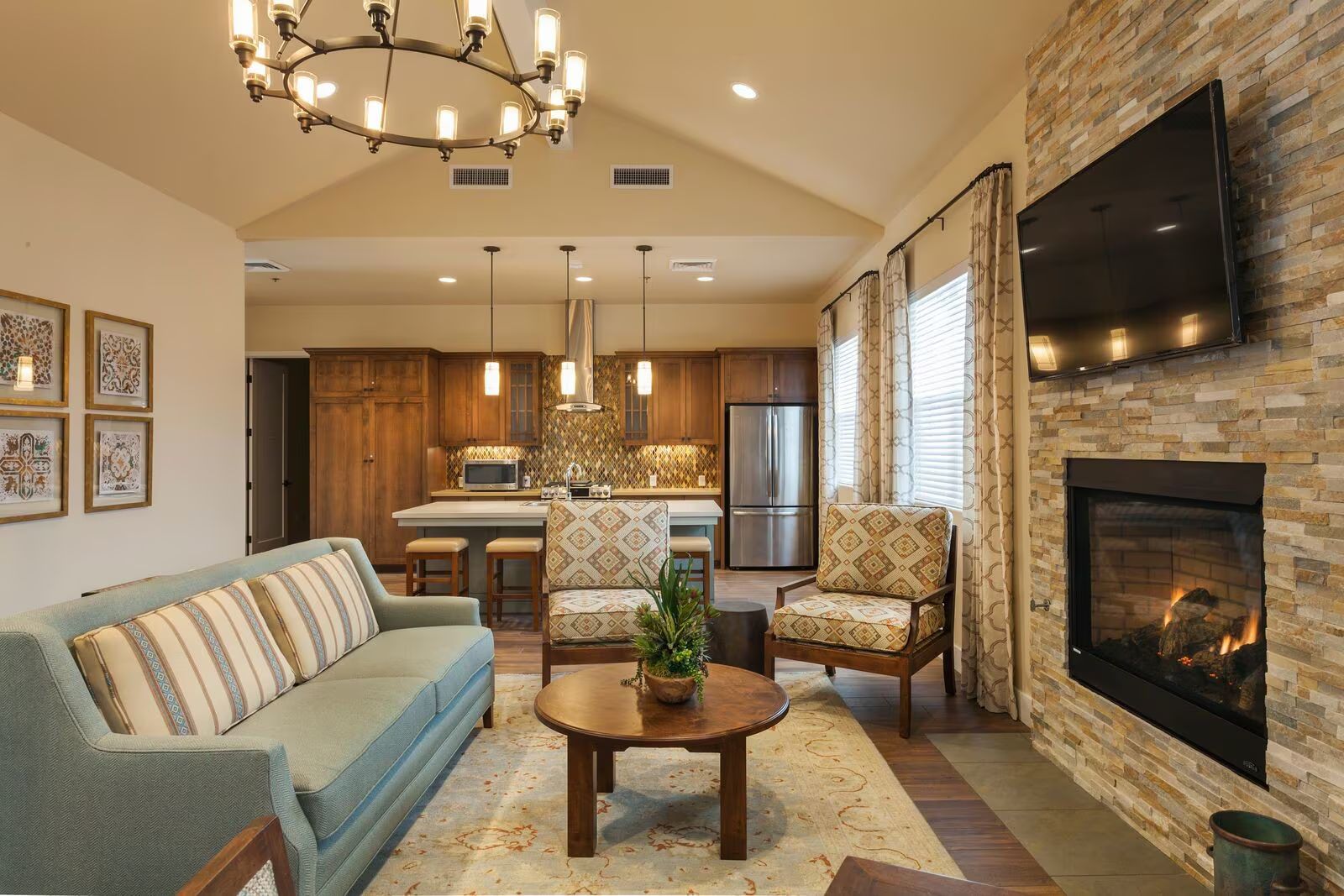 Cozy open-concept living room with stone fireplace, wall-mounted TV, sofa, and kitchen in the background