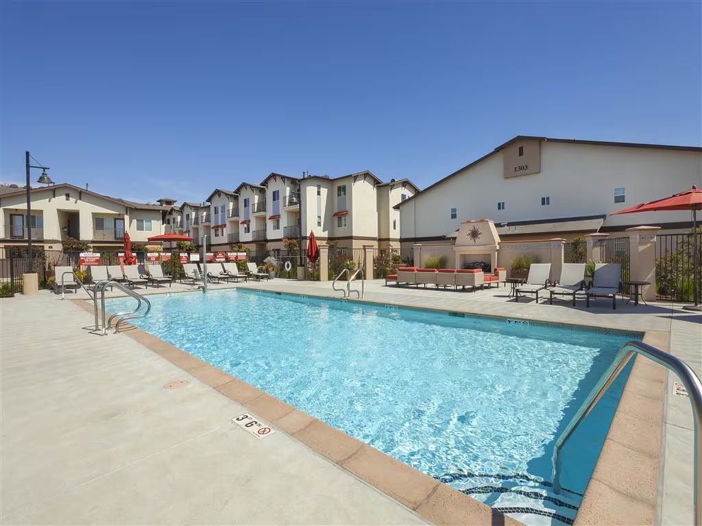Apartment complex pool with blue water, lounge chairs, and surrounding buildings under a clear sky