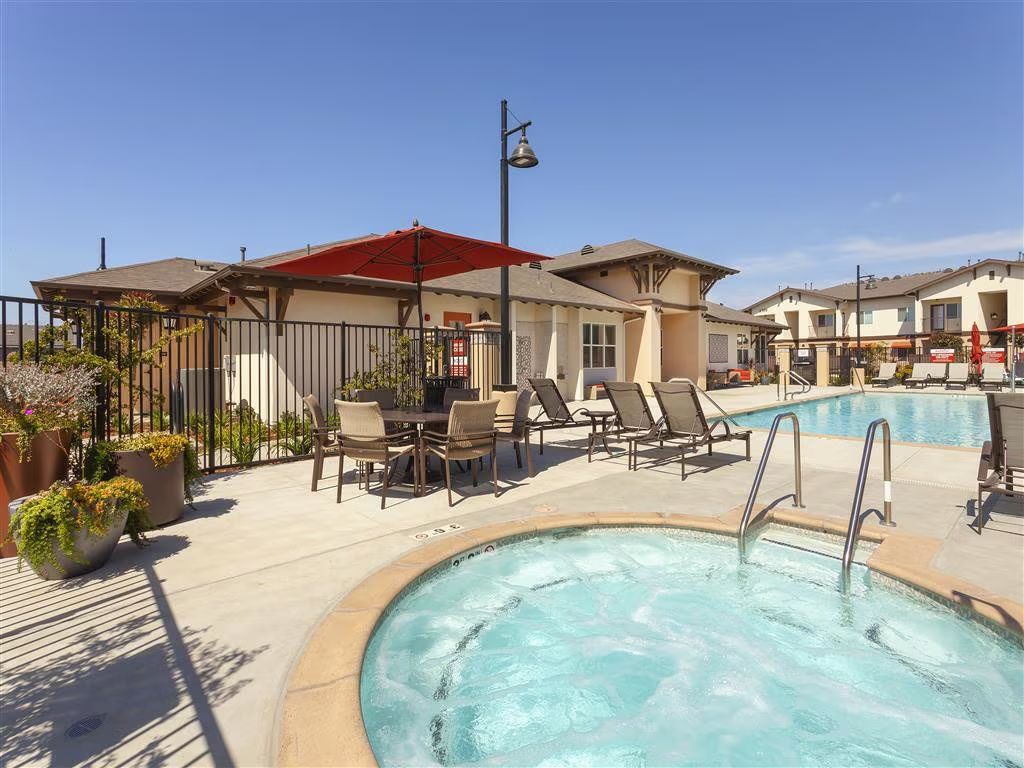 Apartment courtyard with a swimming pool, lounge chairs, and shaded seating under a blue sky