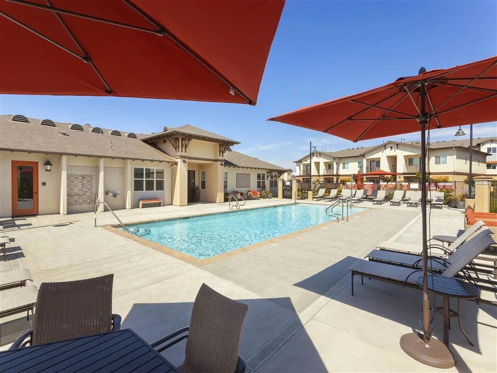 Luxury resort pool with red umbrellas, lounge chairs, and cabanas under a bright blue sky