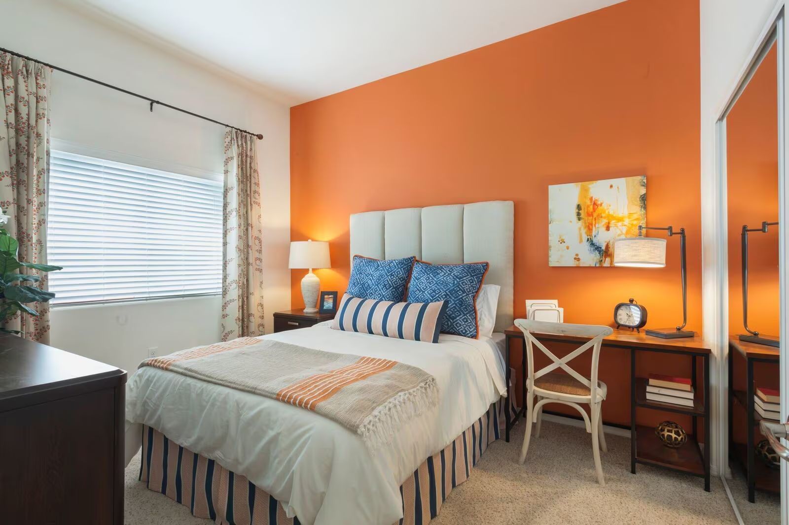 Bright bedroom with orange accent wall, white bed, wooden nightstand, and window blinds