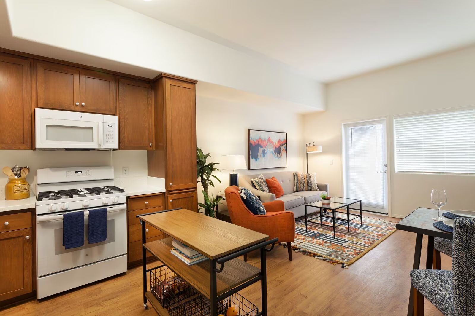 Bright living room with orange sofa, patterned rug, and open kitchen with white appliances and wood cabinets