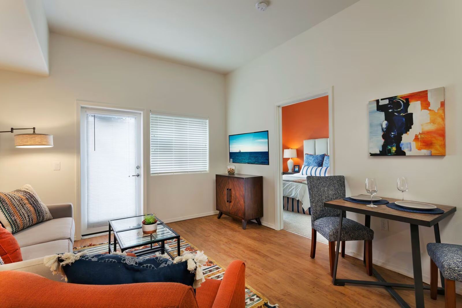 Bright living room with orange chair, coffee table, desk, and colorful wall art near a small TV cabinet