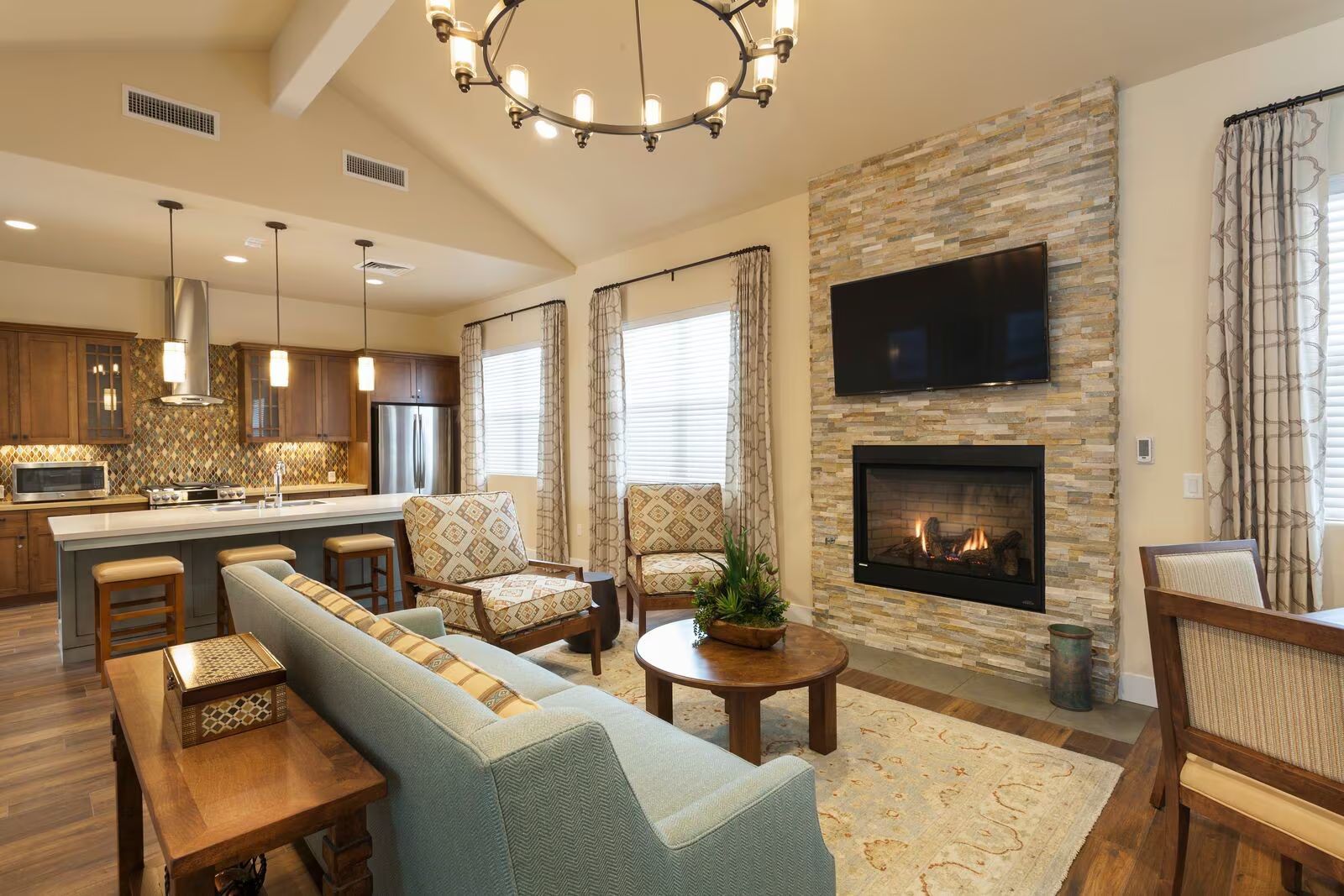 Cozy open-plan living room with stone fireplace, TV, kitchen island, and warm neutral decor