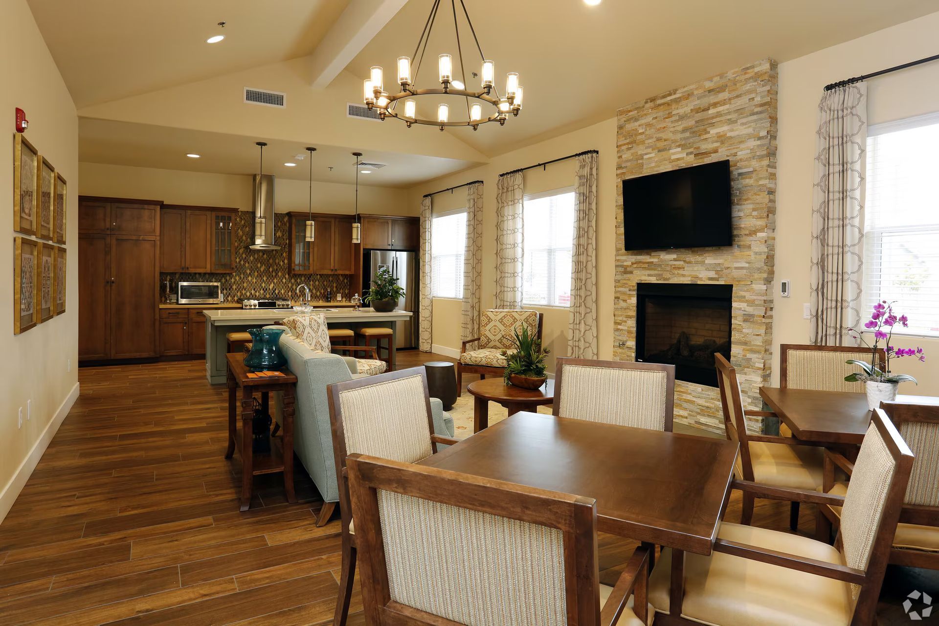 Bright open dining and living room with wooden table, stone fireplace, kitchen, and chandelier lighting