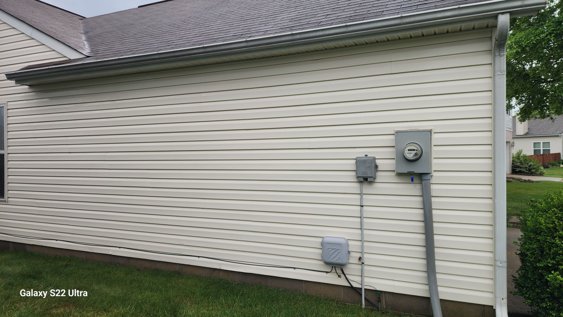 House side with beige siding, utility meters, and a grassy yard along a narrow path