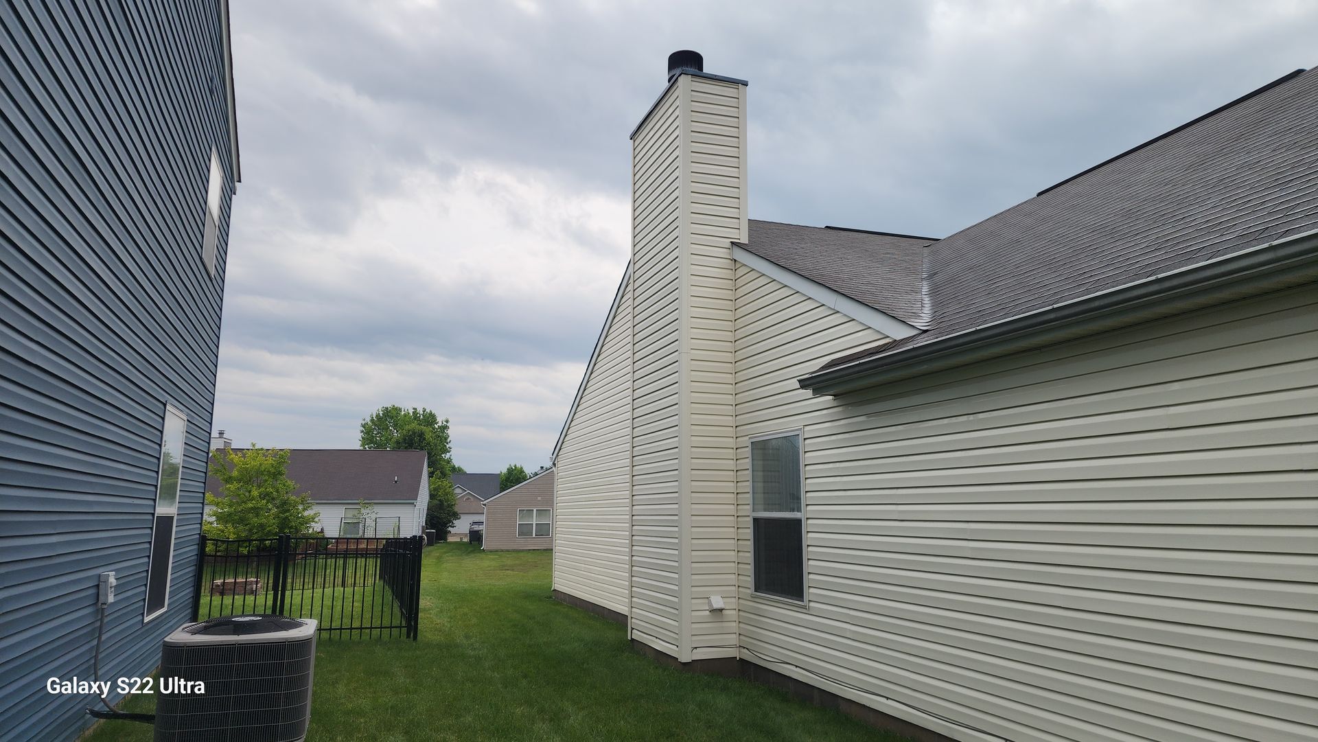 Narrow grassy side yard between white house and black-sided building under cloudy sky