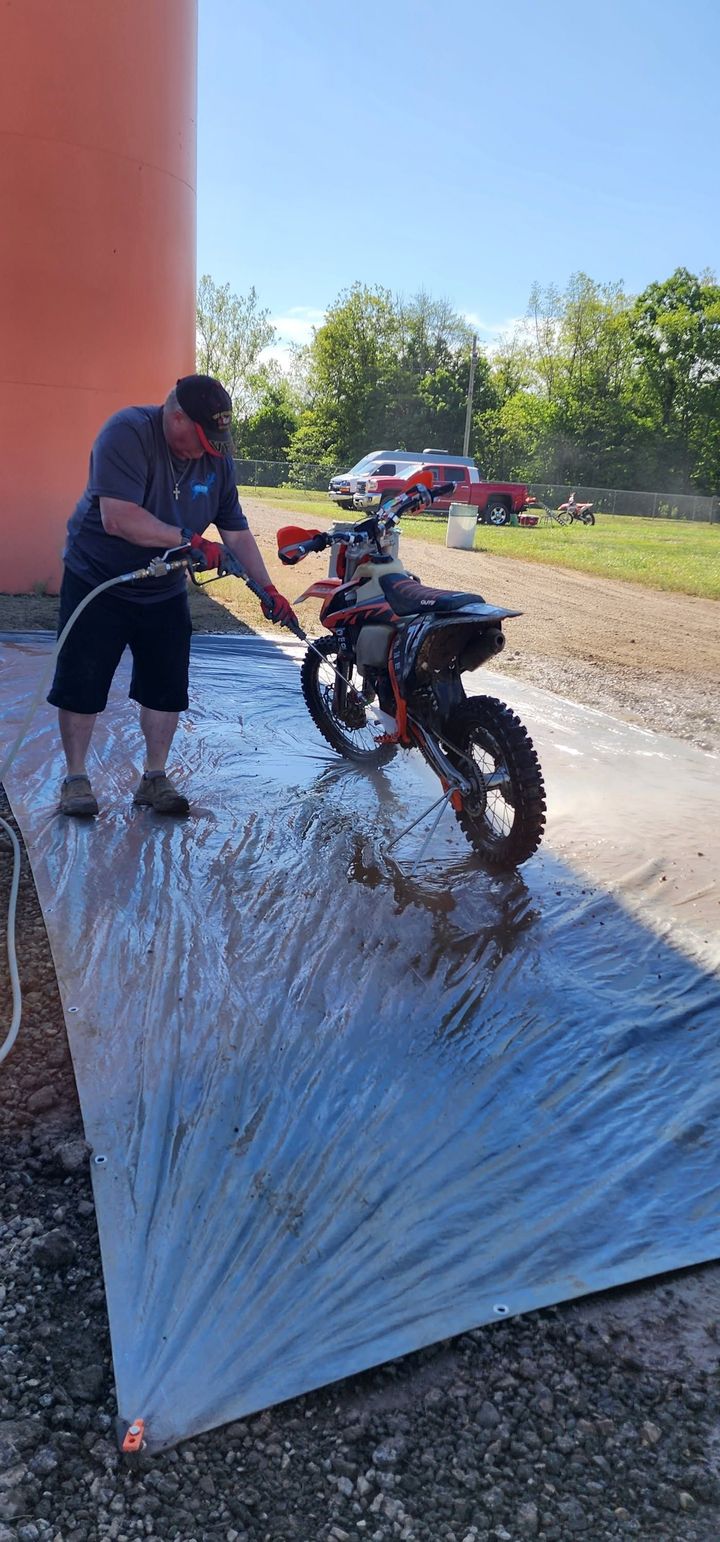 Person washing a black motorcycle on a wet tarp outdoors in bright sunlight.