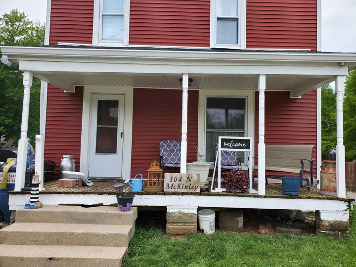 Red house porch with white columns, chairs, and a small sign on a raised wooden deck.