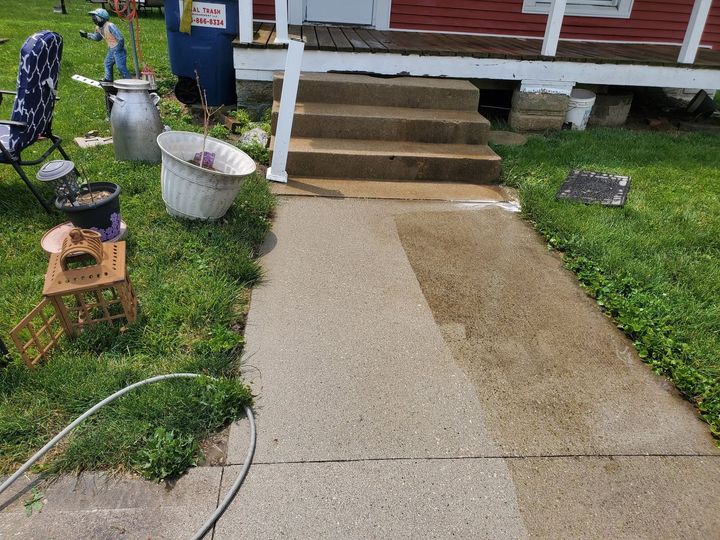 Concrete walkway leading to porch steps in a yard, with flowers and garden items nearby.