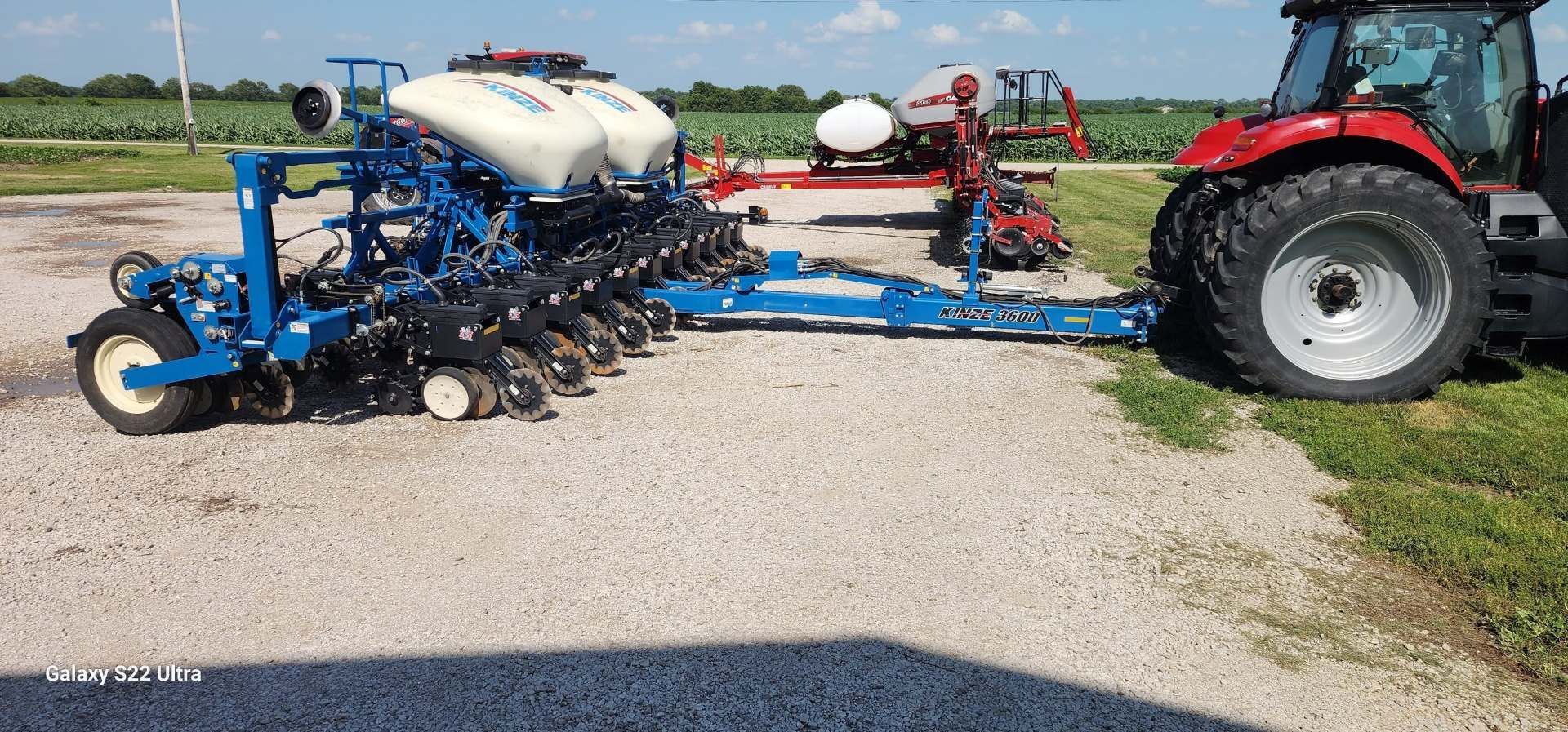 Blue farm planter hitched to a red tractor on a gravel lot, with seed tanks visible.