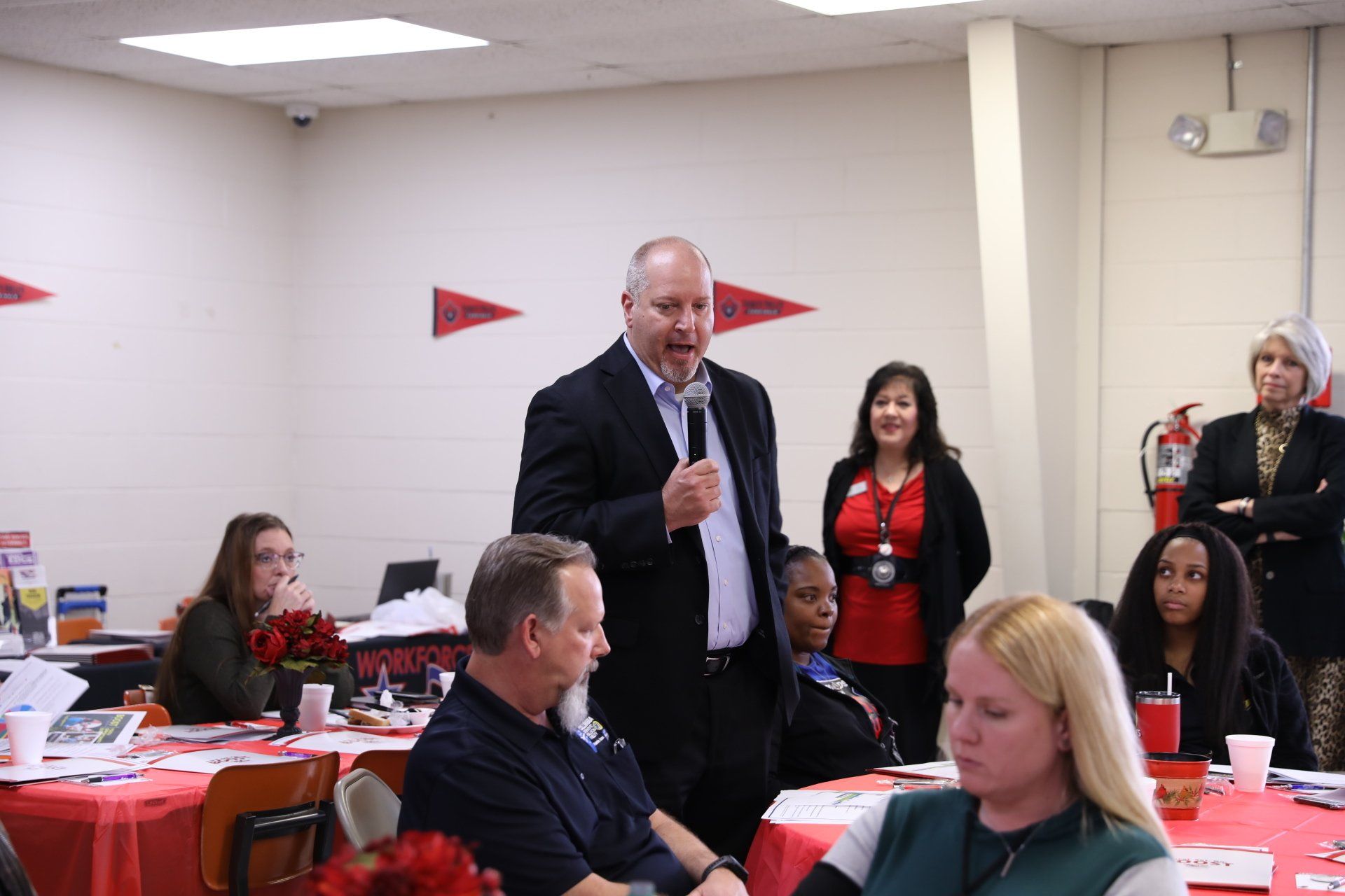 A man in a suit is standing in front of a microphone in a room with people sitting at tables.