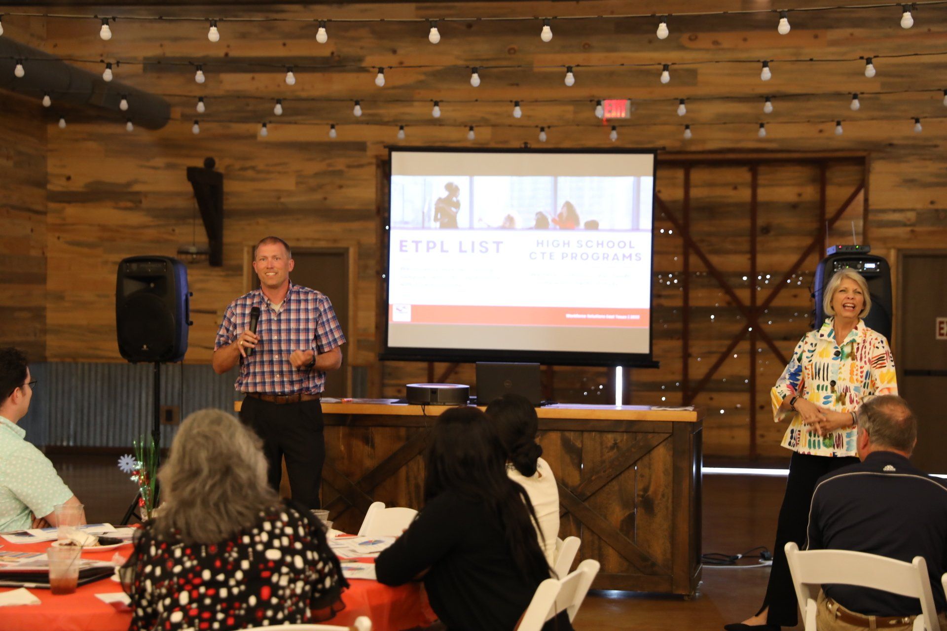 A man and woman are giving a presentation to a group of people sitting at tables.