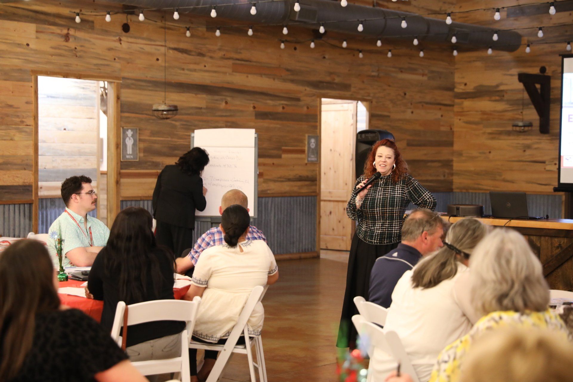 A woman is giving a presentation to a group of people sitting at tables in a room.