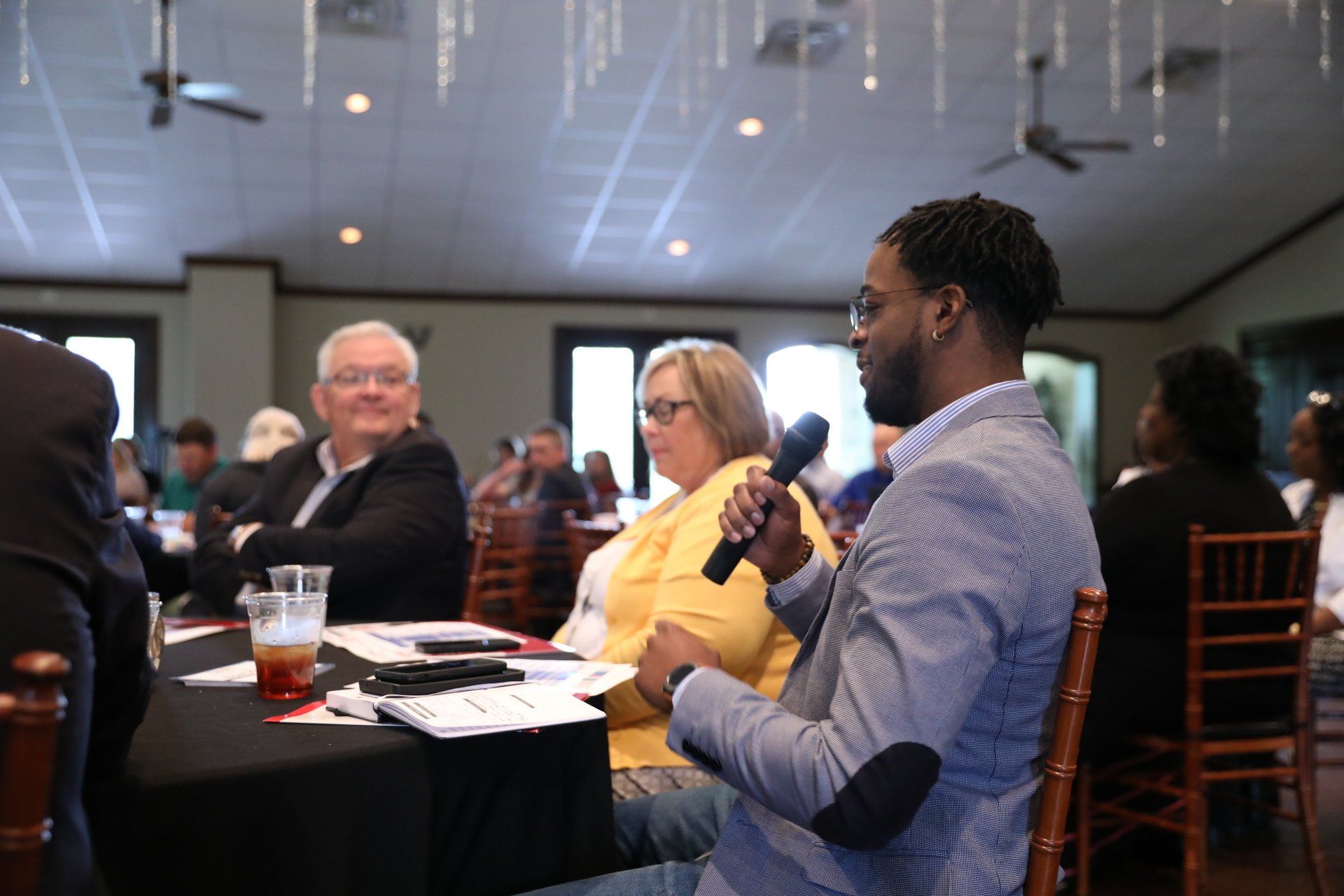 A man is holding a microphone while sitting at a table with other people.