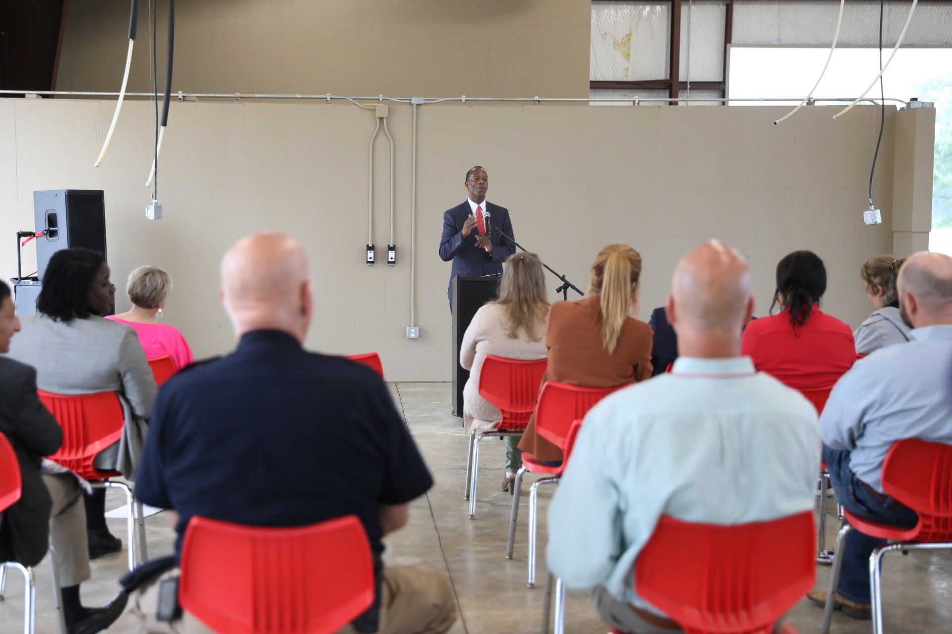 A man is giving a speech to a group of people sitting in red chairs.