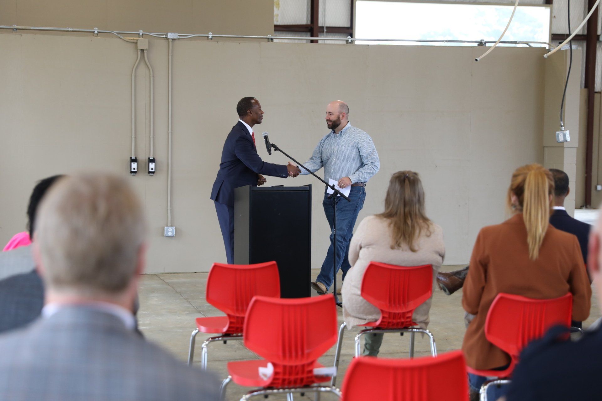 A man shaking hands with another man at a podium in front of a crowd of people sitting in red chairs.