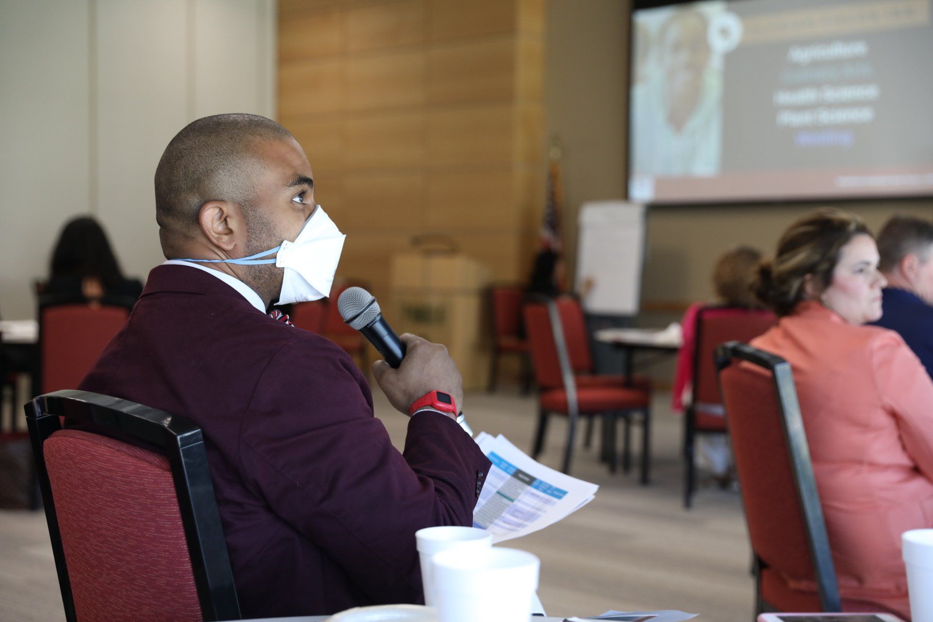 A man wearing a mask is sitting at a table holding a microphone.