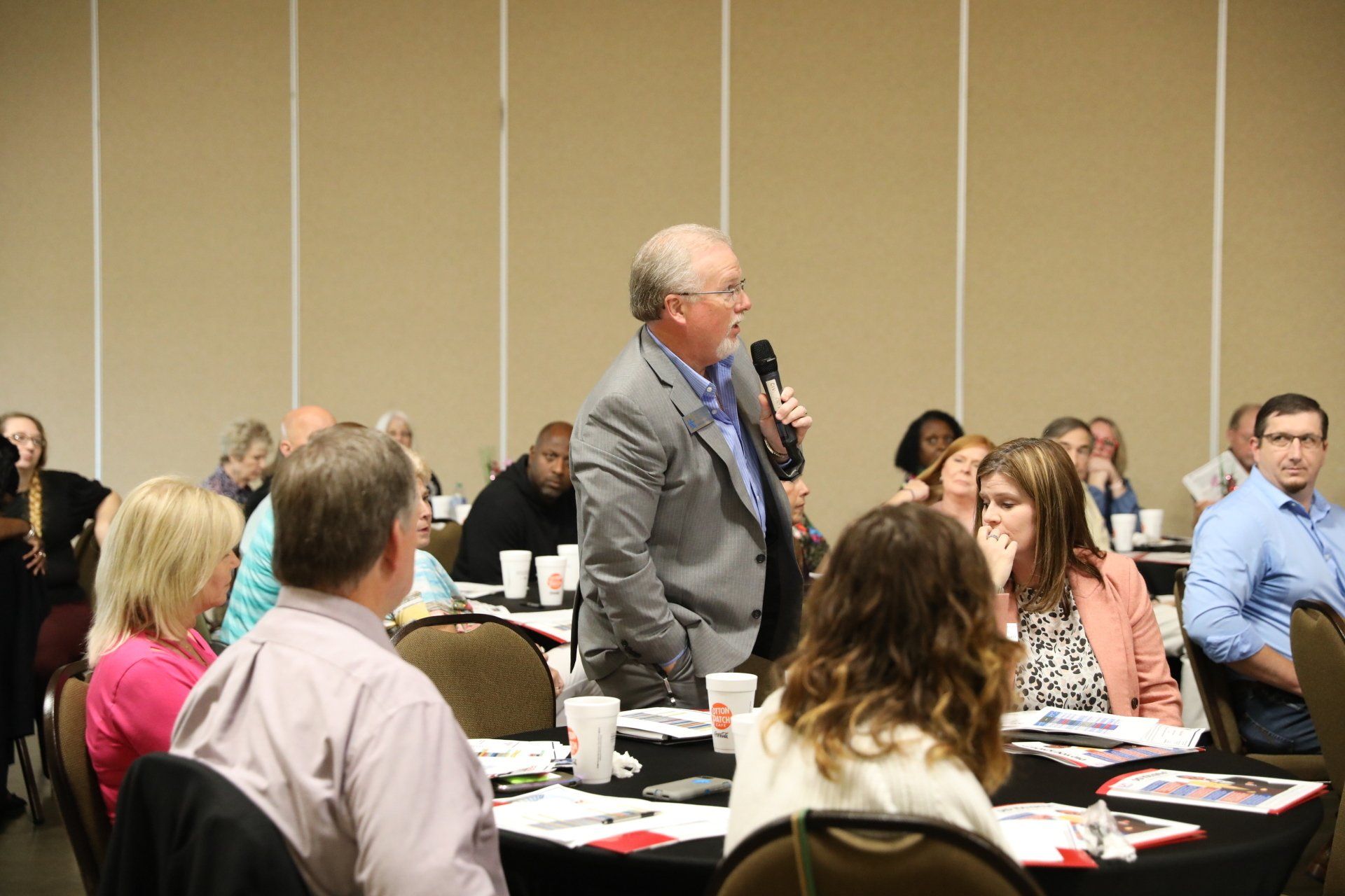 A man is holding a microphone and talking to a group of people sitting at tables.
