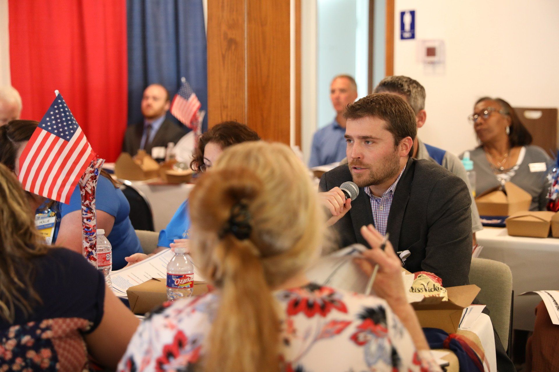 A man is talking into a microphone while sitting at a table with other people.