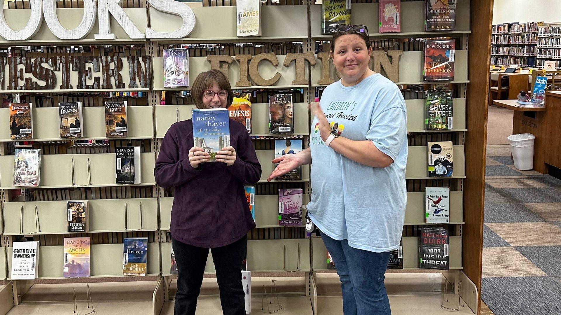 Two women are standing in front of a bookshelf in a library holding books.
