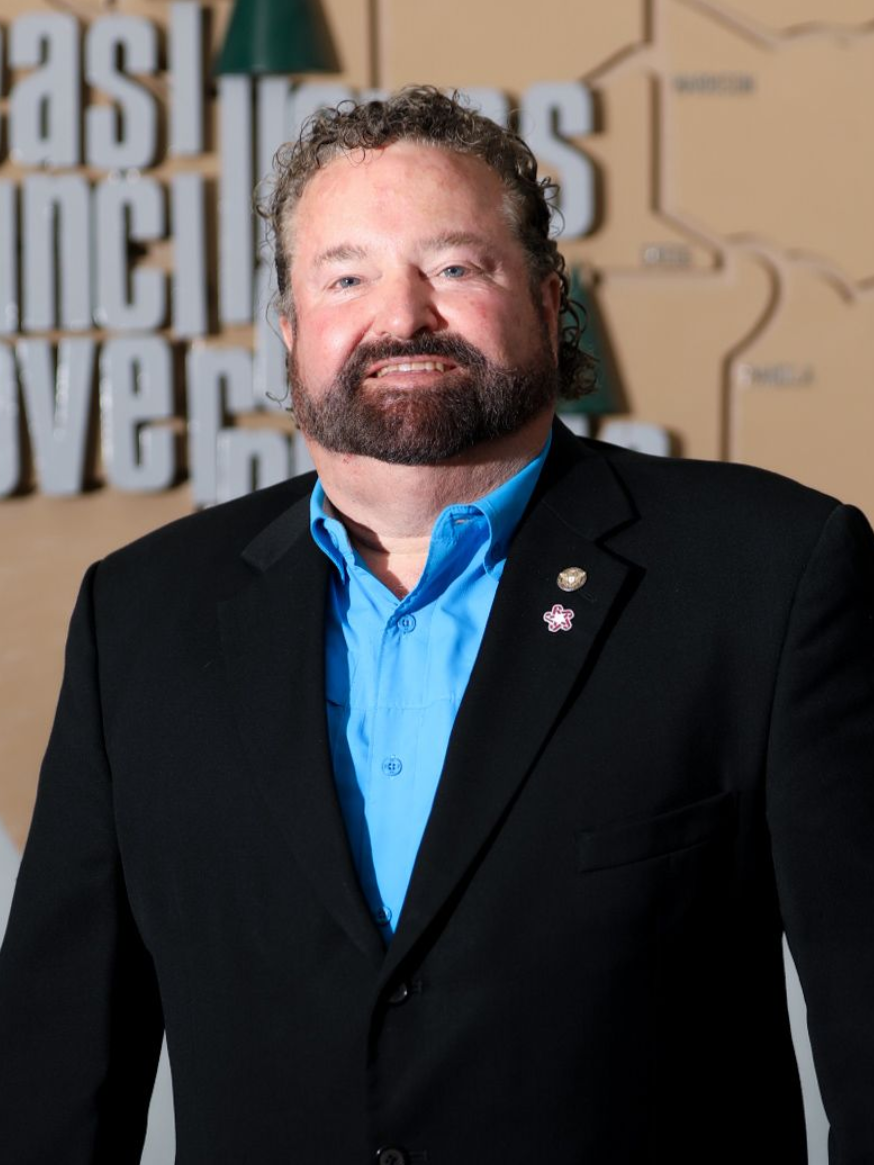 A man in a black suit and bright blue shirt stands smiling at a formal event backdrop.