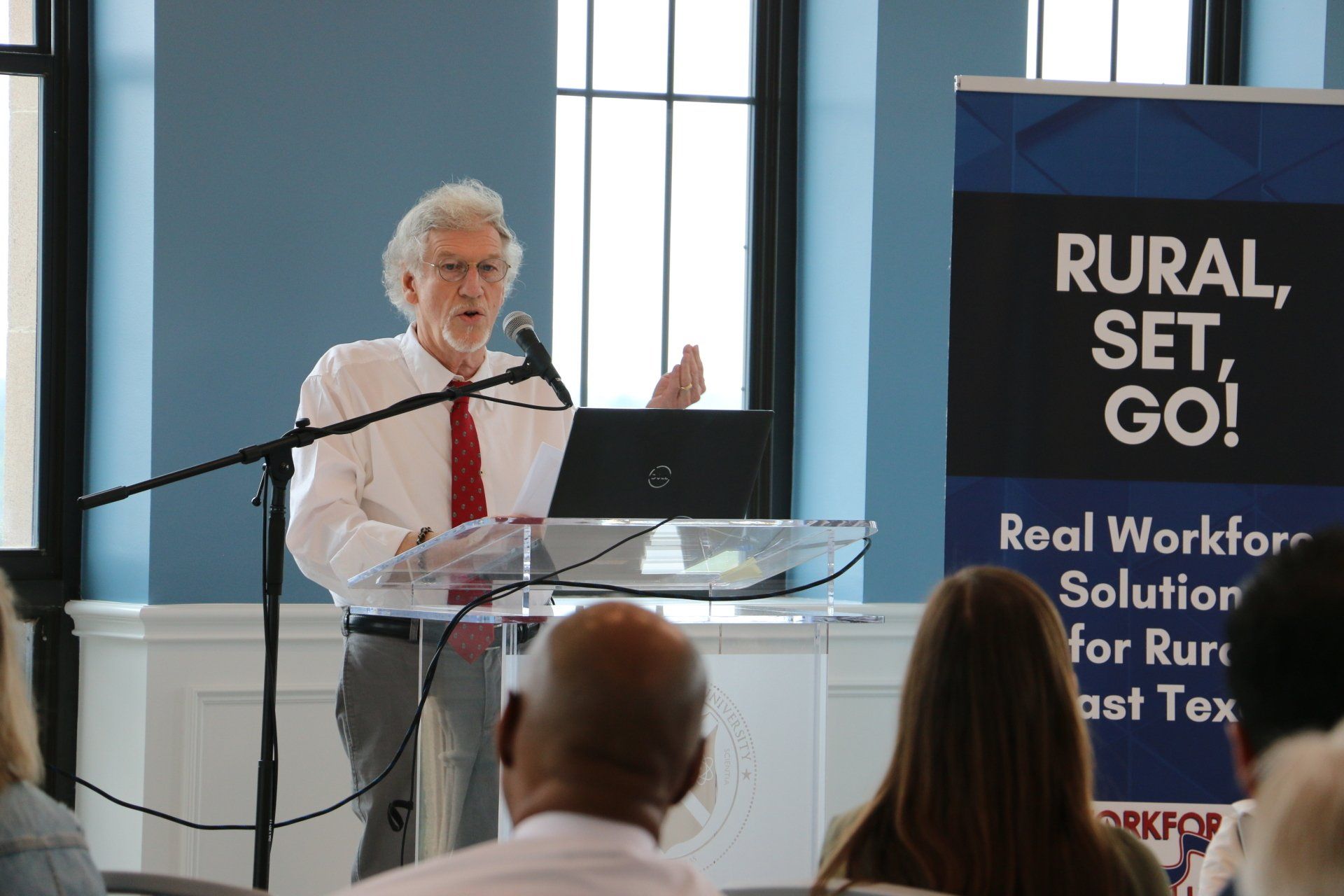 A man is giving a speech at a podium with a sign behind him that says rural set go