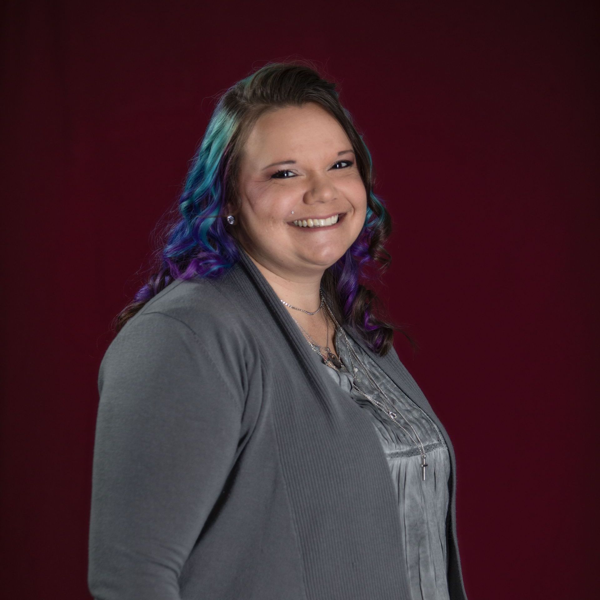 Smiling person with blue-purple hair in a gray top against a dark red studio backdrop