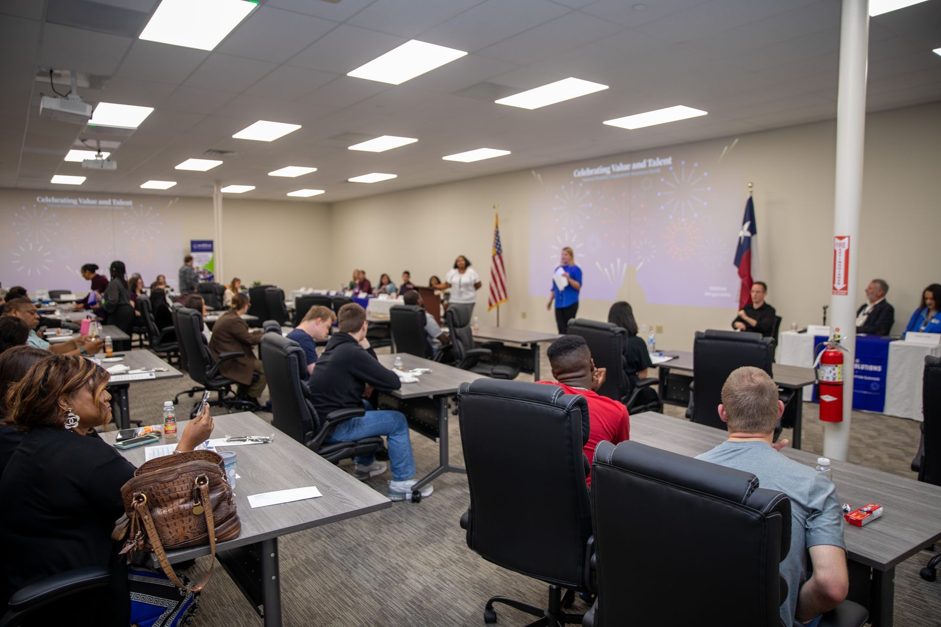 Community meeting in a conference room, with people seated facing a speaker near flags and a screen.