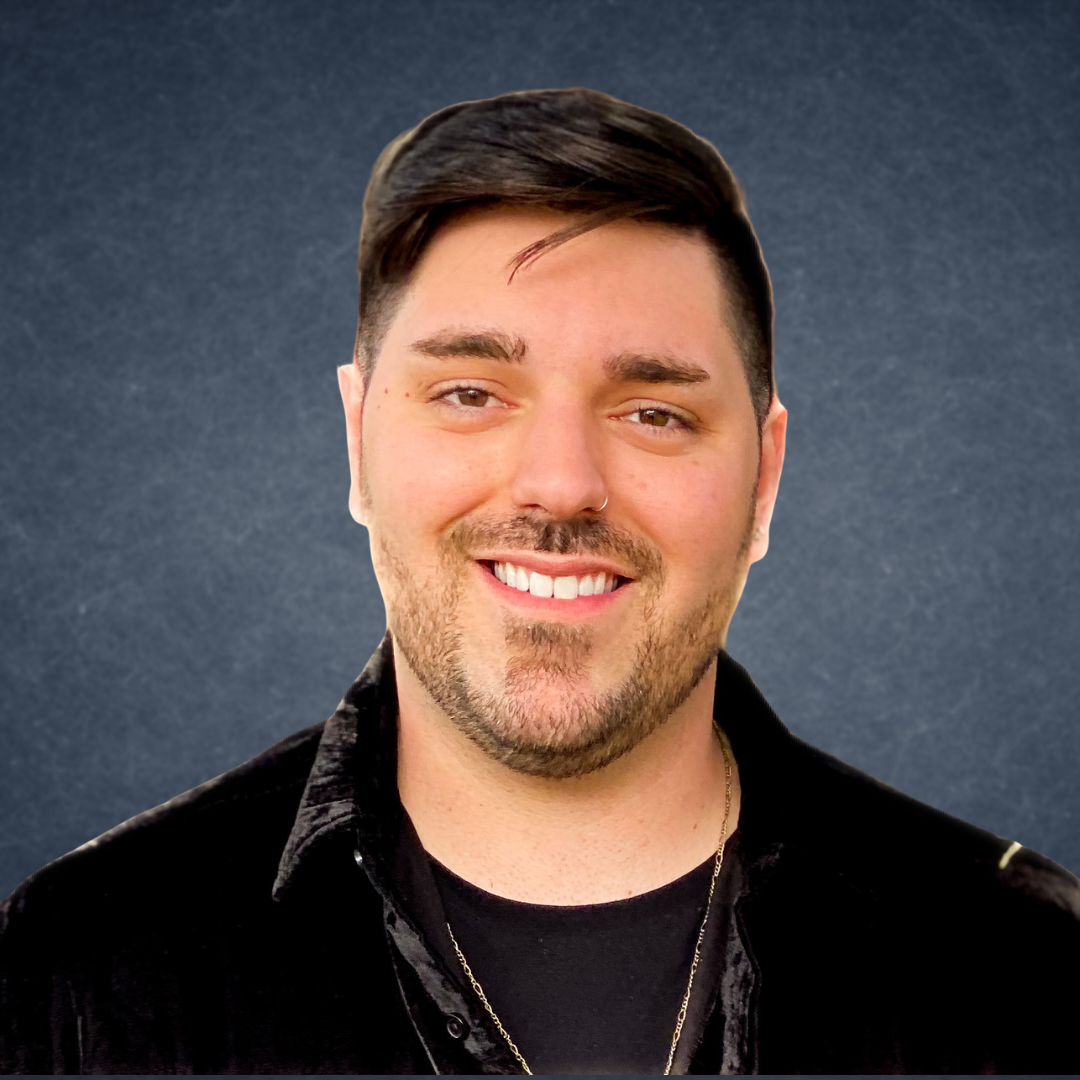 Smiling person with short dark hair and beard, wearing a black jacket against a gray studio background