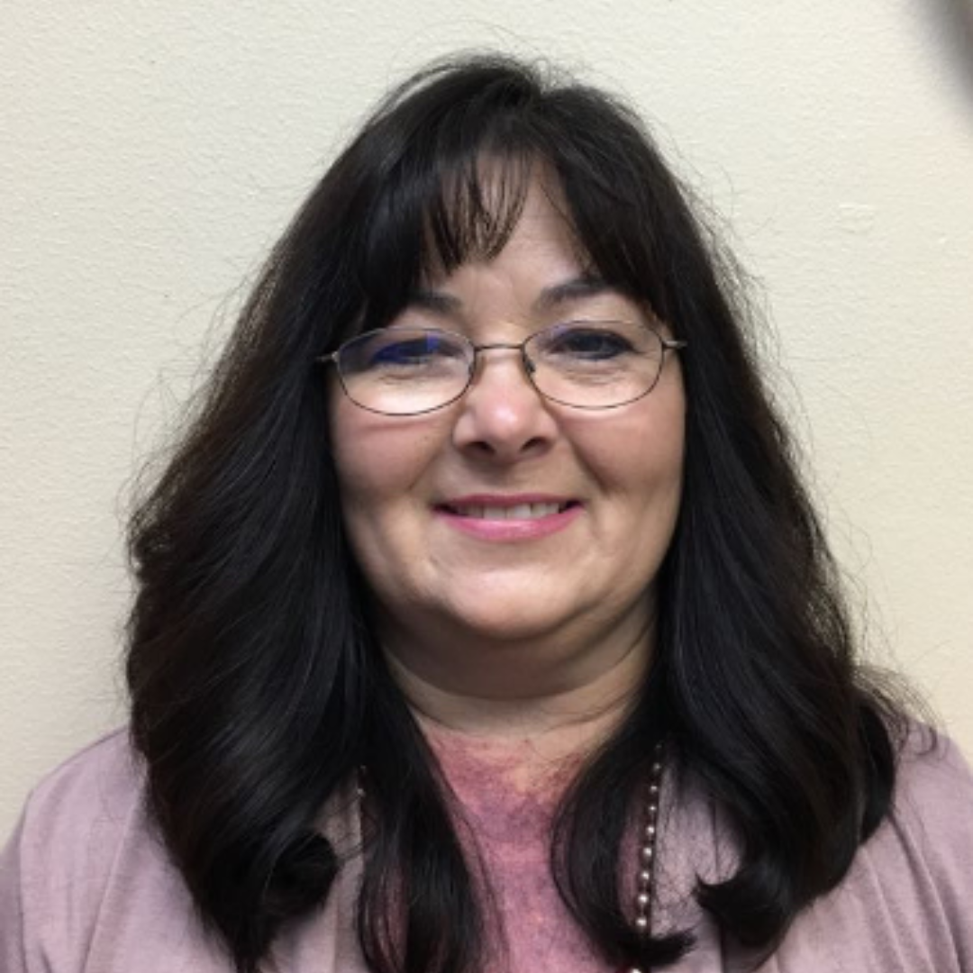 Smiling woman with glasses and long dark hair against a light wall, wearing a pink top