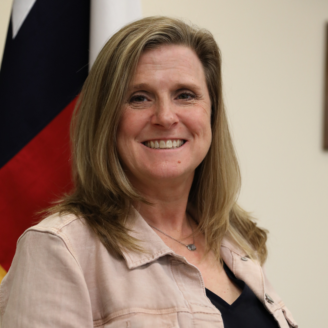 Smiling woman in a light pink jacket standing indoors near a flag