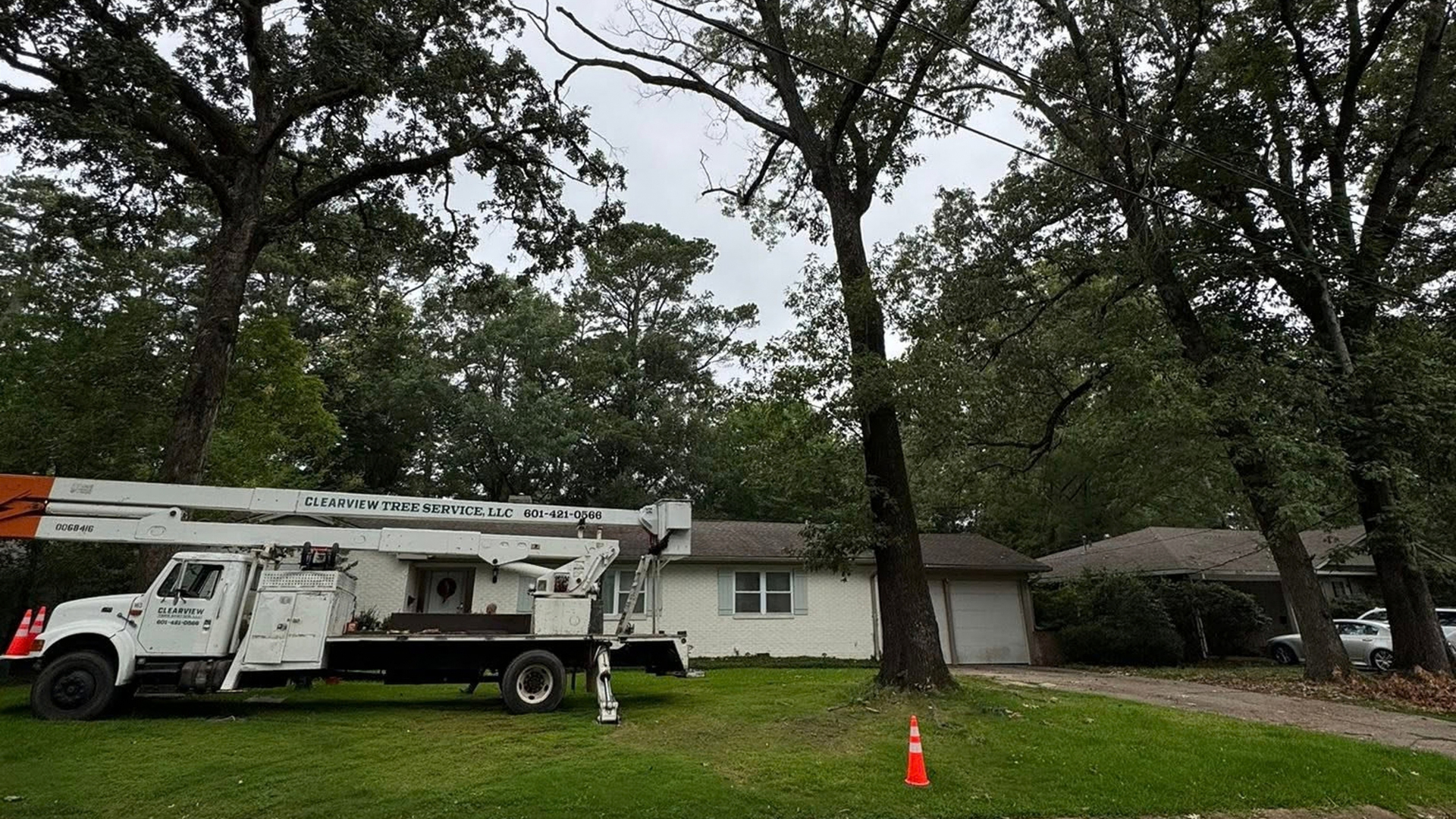 Utility truck and bucket lift parked in front of a house with trees and an orange cone on the lawn