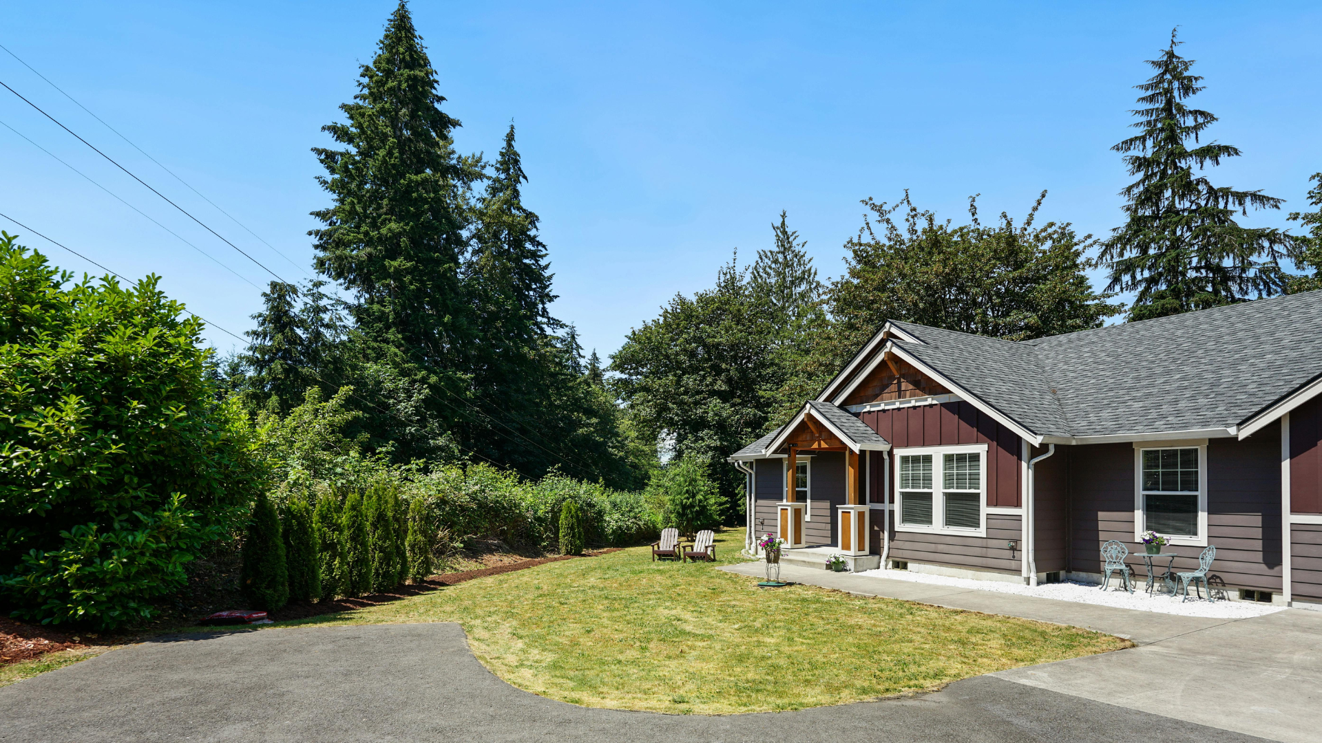 Suburban house with red trim, gravel driveway, and tall evergreen trees under a clear blue sky
