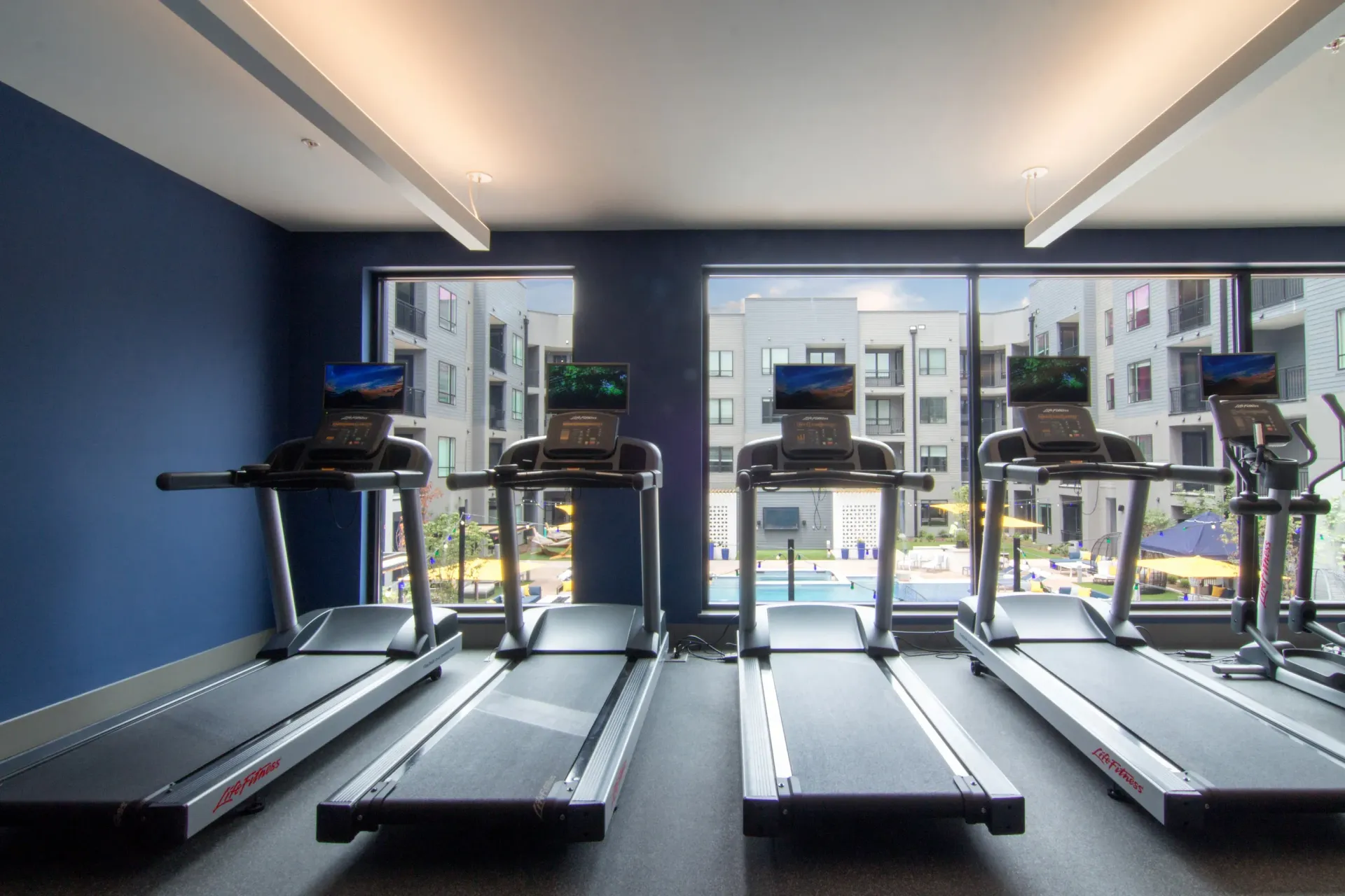 Gym interior with four treadmills lined up by large windows overlooking the outdoor pool at Chroma, offers STL downtown apartments.