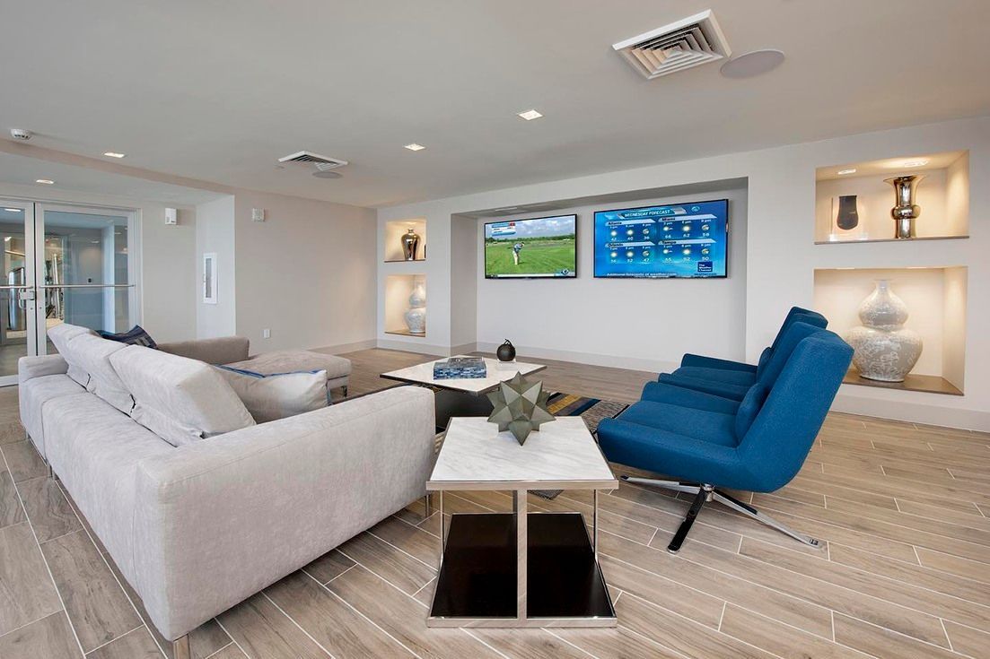 Modern resident lounge with gray sofa, blue chairs, and wall-mounted TVs at Chroma, offers apartments near UMSL Campus.